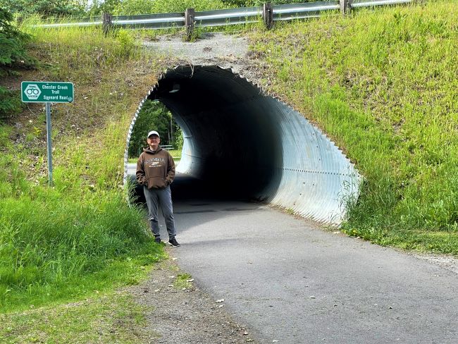 Person standing by a large metal tunnel under a grassy embankment with a green sign nearby.