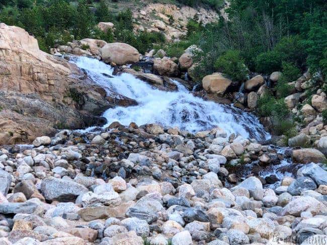 a scenic view of a waterfall cascading down rocky terrain, forming an alluvial fan. The waterfall is surrounded by large, rounded boulders and smaller stones of varying sizes. The water appears to flow gently, creating a white frothy texture as it streams over the rocks. In the background, dense greenery, including trees and shrubs, lines the edges of the rocky area. The landscape is natural and untouched, illustrating the beauty of nature in its raw form.