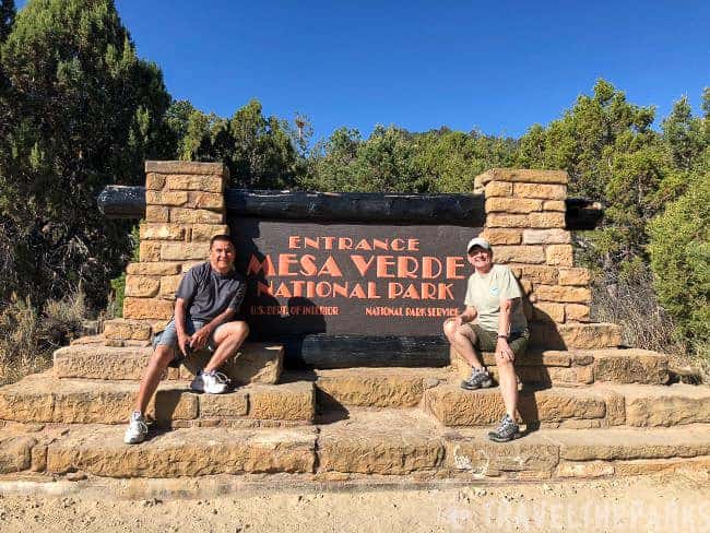 Two people sitting in front of the Mesa Verde National Park entrance sign.