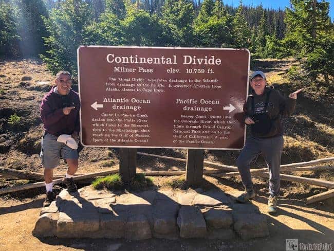 Milner's Pass Two people stand beside a sign indicating the Continental Divide in Rocky Mountain National Park, surrounded by scenic landscapes.