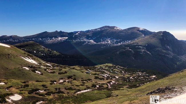 Trail Ridge Road scenic view of the Rocky Mountains from a hillside, showcasing majestic peaks and lush greenery in the foreground.