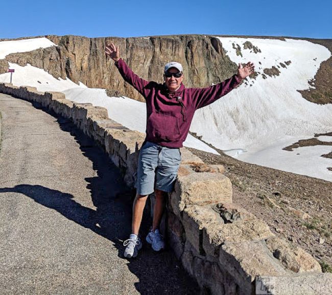 A man stands on a stone wall in Rocky Mountain National Park, arms outstretched, embracing the breathtaking scenery around the Lava Cliff, Trail Ridge Road