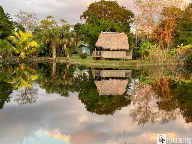 Landscape with reflective water, palm trees, and a thatched-roof chickee hut at sunset.