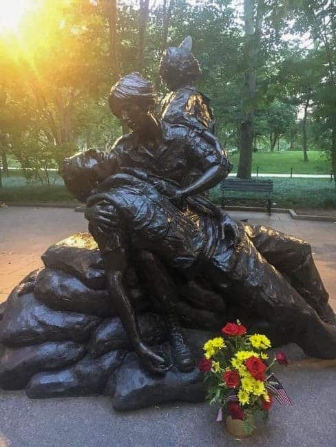 Vietnam Women’s Memorial bronze statue of a nurse holding a wounded soldier with another soldier kneeling behind her; flowers in foreground.