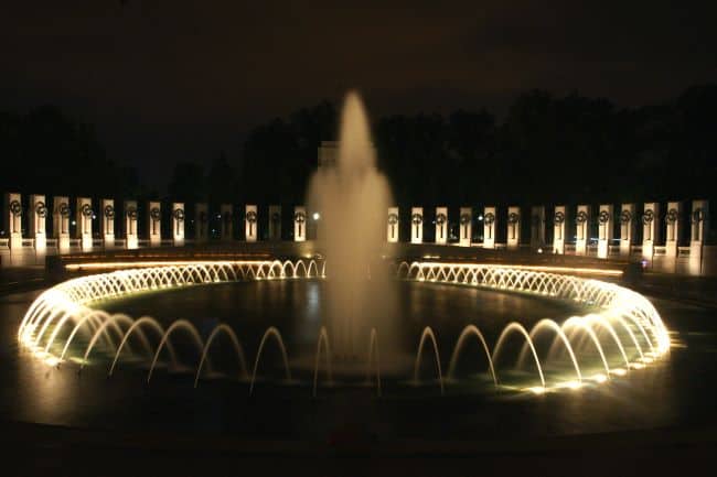 World War II Memorial-A circular illuminated fountain at night with arched water jets and background columns.