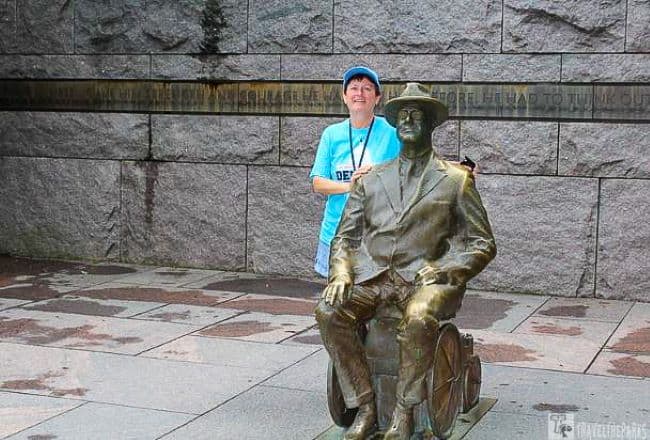 Bronze statue of Franklin D. Roosevelt in a wheelchair with a person standing beside it in front of a stone wall.