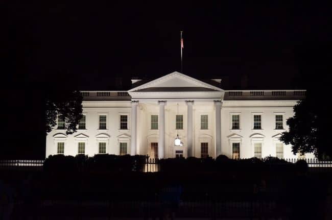 The White House illuminated at night with a flag on the roof and trees on either side.