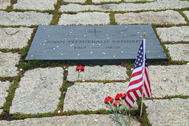 Arlington National Cemetery Gravestone of John Fitzgerald Kennedy with coins, red carnations, and an American flag.