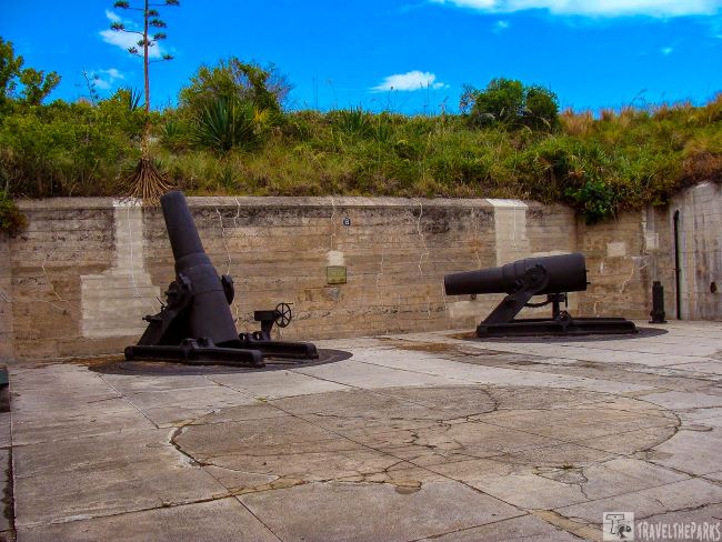 Two historical cannons on concrete mounts at Fort DeSoto Beach with a wall and greenery in the background.