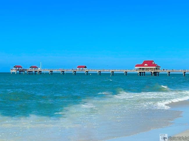 Pier with red-roofed structures extends over turquoise waters at Clearwater Beach under a clear blue sky.