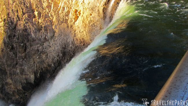 Brink of the Falls-Grand Canyon of Yellowstone-A waterfall flows over a rough, multicolored rock cliff into a moving body of green water.