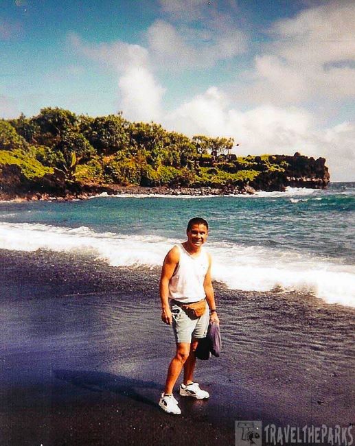 Honokalani Black Sand Beach" Person standing on a black sand beach with ocean waves and a green cliff in the background.