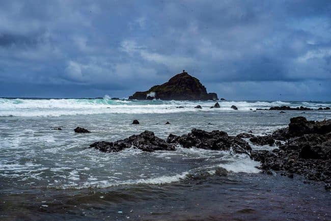 Koki Beach-Alau Island: Rocky coastline with waves crashing onto dark rocks and an overcast sky above.