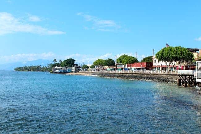 a scenic coastal view of Lahaina Maui with a vibrant blue ocean in the foreground. A stone seawall runs alongside the water, leading to a small coastal town. A row of buildings painted in various colors such as red, yellow, and beige lines the shore. Lush green trees are evenly spaced among the buildings, adding a touch of nature to the scene. The sky is clear with a few scattered clouds, and the distant hills to the left add depth to the background.