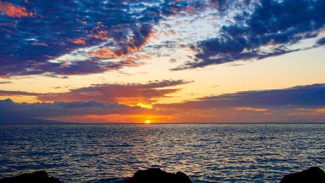 a scenic view of a sunset over the ocean, likely on the island of Maui. The sun is partially visible near the horizon, casting a warm golden light across the water. The sky is filled with an array of clouds, ranging from dark grey to reddish-orange, spreading across the top of the image. The ocean appears calm with gentle waves reflecting the hues of the sunset.