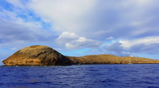 the Molokini Crater located off the coast of Maui, Hawaii. The scene features a crescent-shaped, partially submerged volcanic crater rising from the ocean. The crater exhibits a rugged, rocky surface characterized by shades of brown and light tan, contrasting against the expansive deep blue ocean surrounding it. Sparse vegetation can be seen in some areas of the crater. The sky above is partly cloudy, with soft white clouds diffusing the sunlight, creating a serene ambiance. A small white sailboat is visible on the right side of the image, providing a sense of scale.