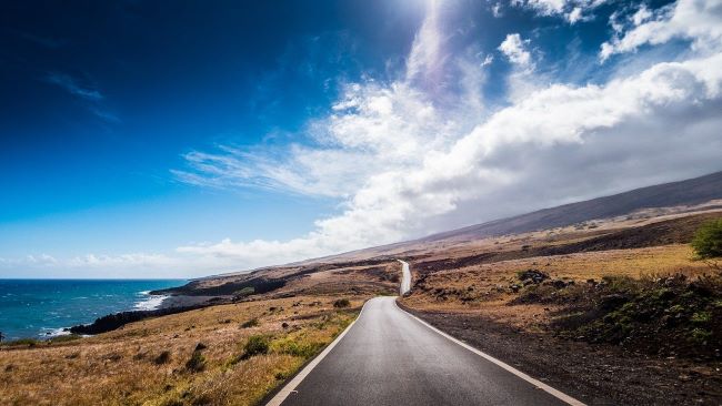 A scenic Piilani Highway leads toward distant hills under a bright blue sky with white clouds.