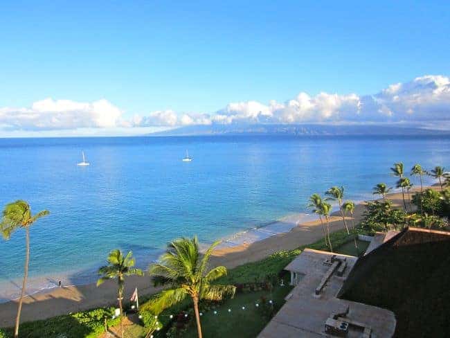 View of Ka'anapali beach in Maui with palm trees and sailboats, with an island and clouds in the distance.