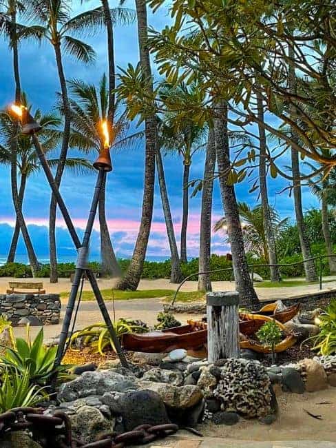 Mammas Fish House-A Hawaiian landscape with lit tiki torches, palm trees, and a colorful sky at sunset.