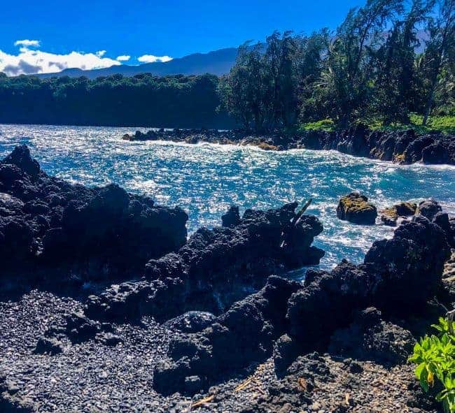 Maui coastline with black volcanic rocks, blue ocean, and dense green vegetation under a clear sky.