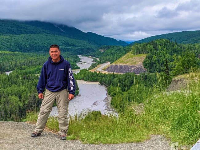 Matanuska River Overlook: Man standing on a grassy overlook with a river and forested hills in the background under an overcast sky.

