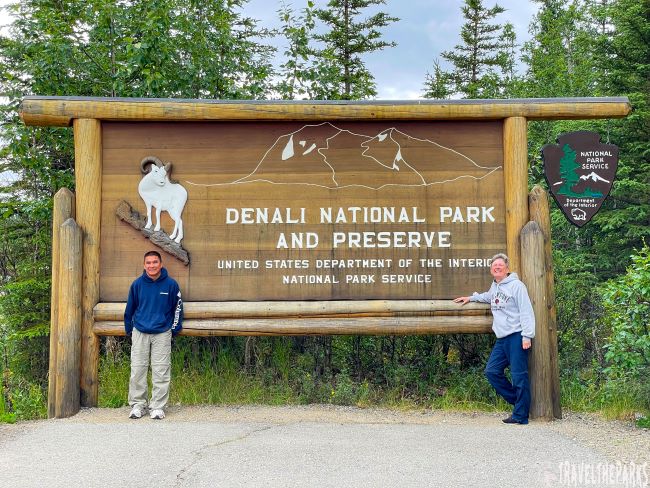 Large wooden sign for Denali National Park and Preserve with carved mountain and Dall sheep, flanked by two people.