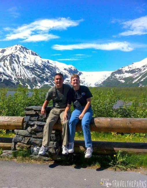 Two people sitting on a wooden fence with snow-covered mountains and Exit Glacier in the background.