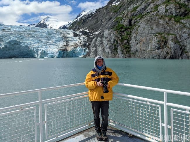 Person in a yellow jacket stands on a deck with Portage Glacier and mountains in the background.