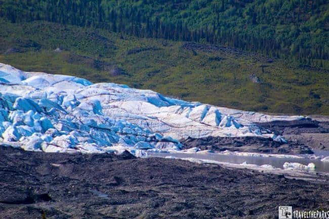 Matanuska Glacier with jagged blue and white ice against dark rocky foreground and green forest background.