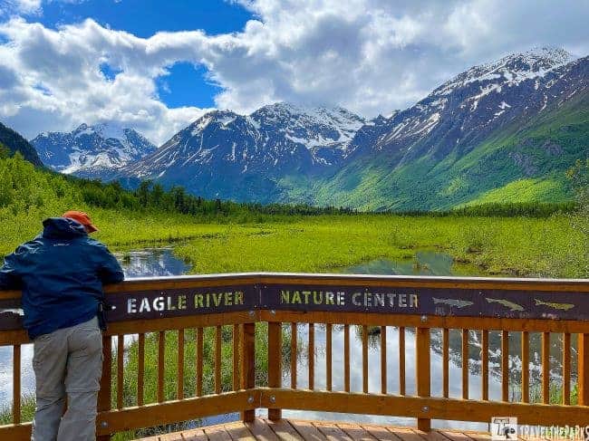 a scenic landscape viewed from a wooden observation deck at the Eagle River Nature Center. In the foreground, a person wearing a blue jacket and beige pants is leaning on the deck's railing, looking out at the view. The railing has an inscription that reads "Eagle River Nature Center". Behind the deck, there is a lush, green wetland area leading to a river that winds through the landscape. Beyond the wetlands, there are towering, snow-capped mountains under a partly cloudy sky. The scene captures the dramatic contrast between the vivid greens of the vegetation and the stark whites and grays of the mountains and clouds.