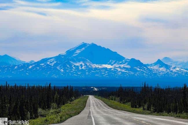 A snow-covered Mt. Drum with a forest and straight road in the foreground under a blue sky.