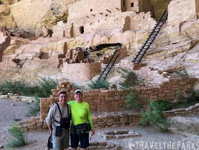 Two people stand in front of an ancient Longhouse cliff dwelling with stone structures and wooden ladders.