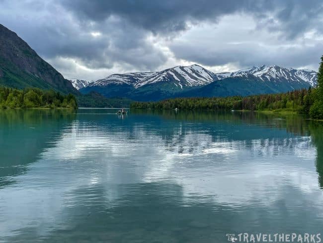 A scenic Kenai lake with a boat, surrounded by green forests and snow-capped mountains under an overcast sky.