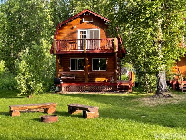 Riverbend Cabins-Fairbanks Alaska-Two-story wooden cabin with a deck in a forested area, surrounded by grass and trees.