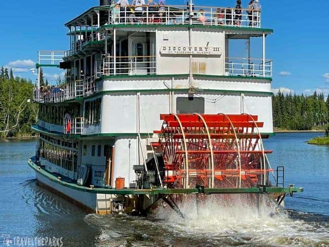 Paddle steamer boat named “Discovery III” with a red paddle wheel, moving through water on the Chena River in Fairbanks, Alaska.
