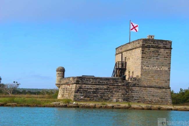 Fort Matanzas, a historic fortification made of coquina stone, located on the edge of a body of water. The fort has a square tower, with a flag flying at the top. The flag is white with a red cross. The tower is fitted with narrow openings and features two visible levels. To the left of the tower, a lower section of the fort displays similar stonework and includes a small dome-like structure. Surrounding the fort are lush green trees and shrubs. The sky above is bright blue with scattered clouds, and the water in the foreground is a clear turquoise, gently lapping against a stone shoreline.