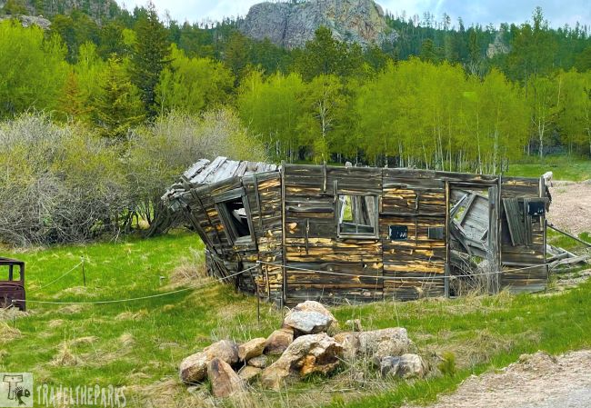 Old, dilapidated wooden structure in a grassy field with trees and a rocky hill in the background.