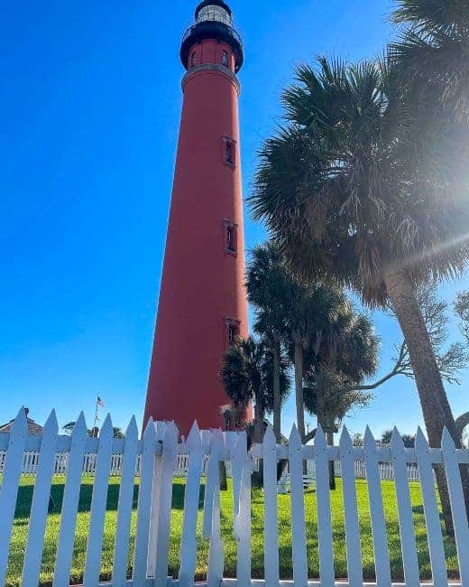 Ponce Inlet Light Station a tall red brick lighthouse with a black railing at the top, under a blue sky, surrounded by palm trees and a white picket fence.