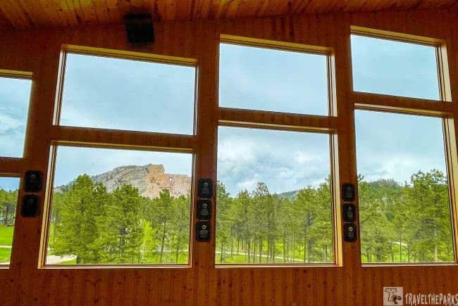 View of Crazy Horse Memorial through large wooden-framed windows with pine trees and an overcast sky.
