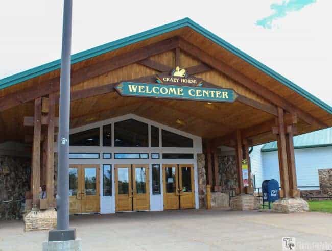 Entrance of the Crazy Horse Memorial Welcome Center with wooden and stone elements, a green shingled roof, and a glass door.