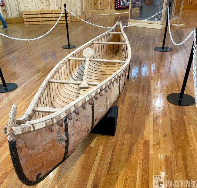 A wooden canoe with a paddle, displayed in a museum on a wooden floor.