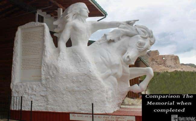 a large white sculpture representing Crazy Horse, mounted on a horse, with a flowing mane and pointed arm. The unfinished real-life Crazy Horse Memorial is visible in the background as a mountain carving. The sculpture is surrounded by a wooden structure. Text is inscribed on a vertical slab that is part of the sculpture.
