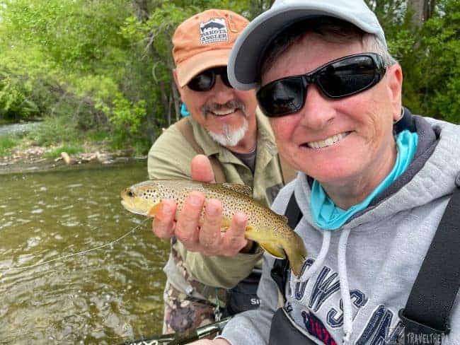 Two people in Rapid Creek, one holding a brown trout, surrounded by greenery in the Black Hills.
