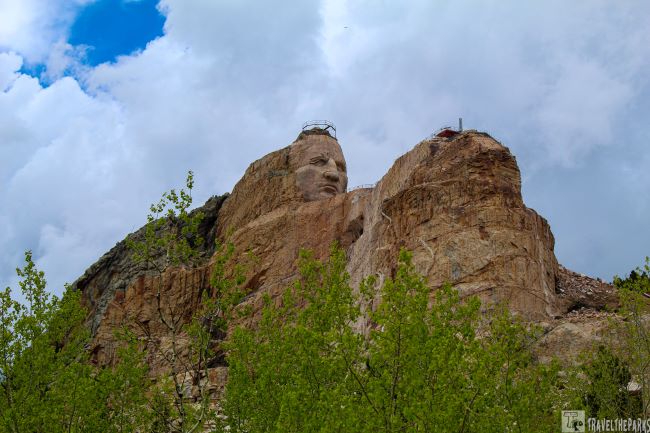 the Crazy Horse Memorial, a massive rock carving in progress. The central feature is a large, detailed sculpture of a face carved into a reddish-brown mountain, with distinct facial features including eyes, nose, and lips. The rock face that forms the sculpture has a rough texture, with varying shades of earth tones, highlighting the natural stone. Perched above the head is scaffold-like metalwork, indicating ongoing construction. To the right, the mountain continues, showing more rough texture and the remnants of chiseling. The sky in the background is overcast with soft, gray clouds, adding to the somber and monumental mood of the scene.
