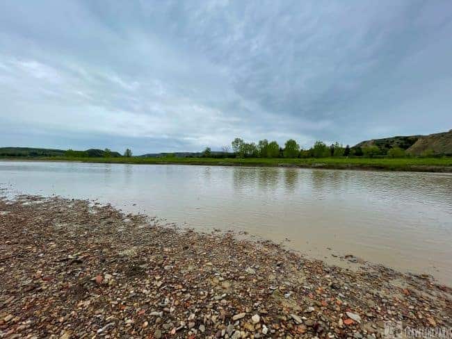 A cloudy sky over a calm Little Missouri river with a rocky shoreline and distant green hills of Theodore Roosevelt National Park.