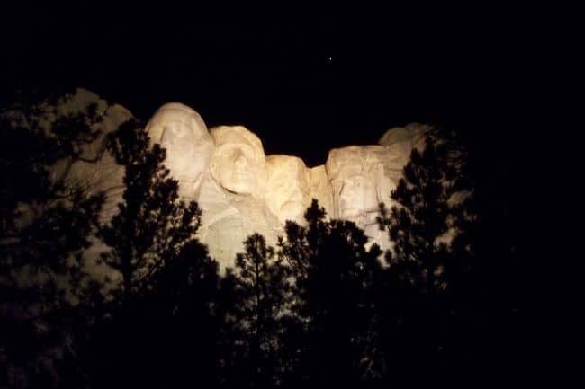 Mount Rushmore lit up at night with pine trees silhouetted in front.