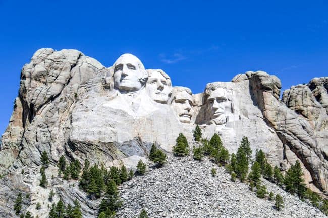 Mt. Rushmore National Monument with clear blue skies and surrounding pine trees.