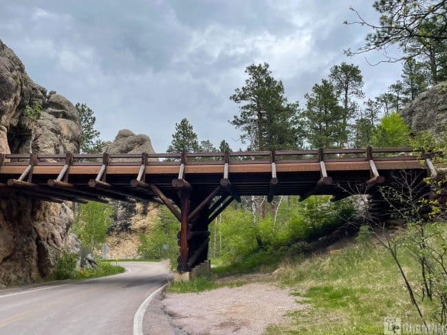 Custer County, SD A wooden Pigtail bridge over a curving road amidst rocky cliffs and trees.


