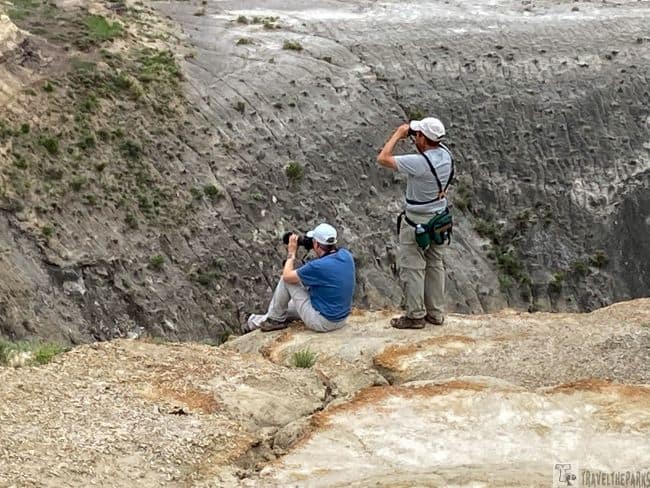 Two people with cameras and binoculars at the edge of a rocky cliff overlooking a canyon.