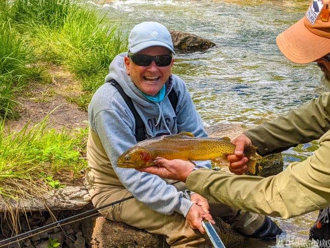 Two people by a stream; one seated, smiling, holding a fishing rod, and the other standing, holding a Cutthroat Trout On Rapid Creek.